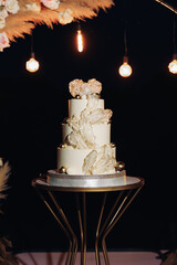 Large luxury wedding cake on a background of a flowering arch in the style of boho. Wedding dessert under the light of evening light bulbs. Wedding decor.