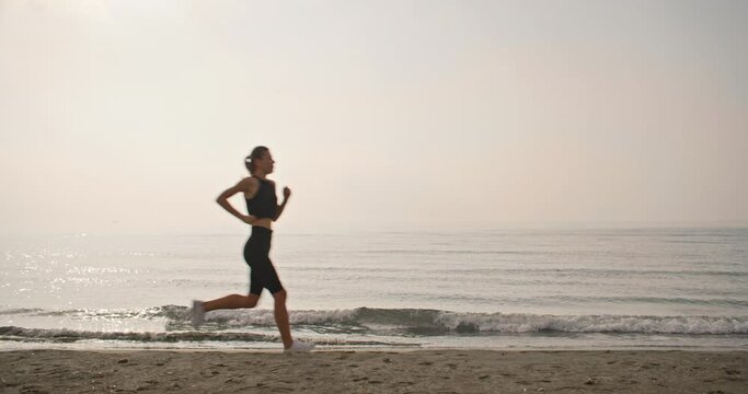 A Caucasian woman on her morning jog runs parallel to the waves of the ocean. Sports and fitness on the sea coast in a beautiful landscape. High quality 4k footage