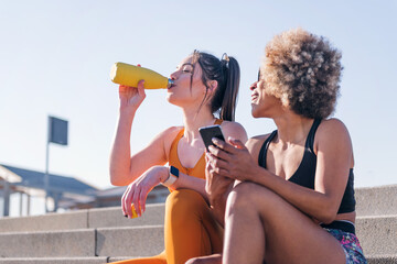 two female runner friends drinking water on a run break while using a mobile sitting on stairs...