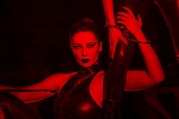 Sexy dominatrix in latex catsuit holding lovers leg in red light - obrazy, fototapety, plakaty