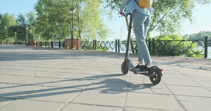 Red-haired young woman kick start rides electric scooter in park near river, green trees, tracking camera, side view.