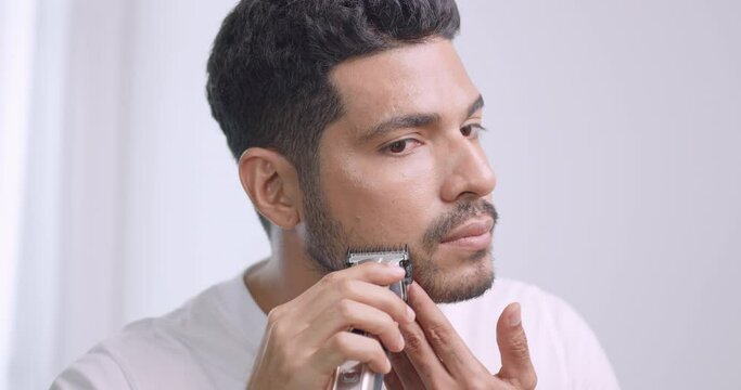 A man is using electric shaver to trim mustache and beard while standing in front of mirror in bathroom at home.