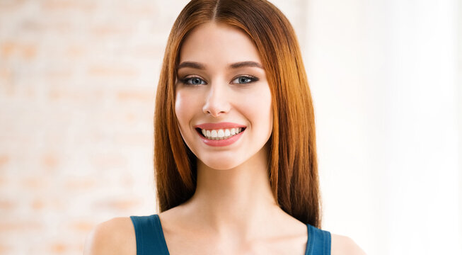 Portrait image of toothy smiling happy woman standing near window, indoors. Brown hair girl at optimistic, positive or dental health care, stomatology concept.