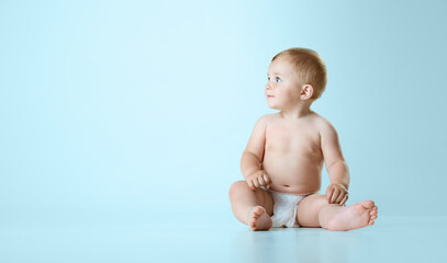 Beautiful little baby, toddler in diaper sitting calmly on floor and looking away against light blue studio background. Concept of childhood, newborn lifestyle, happiness, care. Copy space for ad - obrazy, fototapety, plakaty