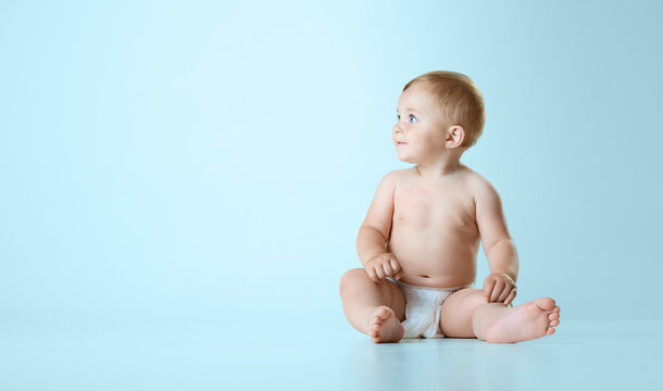 Beautiful little baby, toddler in diaper sitting calmly on floor and looking away against light blue studio background. Concept of childhood, newborn lifestyle, happiness, care. Copy space for ad
