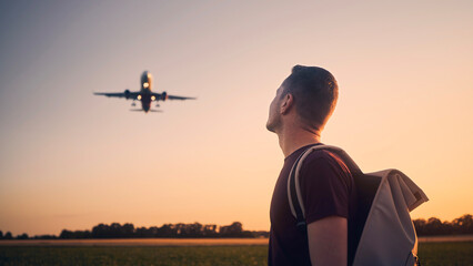 Man with backpack looking up to airplane landing at airport during beautiful summer sunset. .