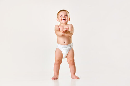 Portrait of little baby girl, toddler in diaper standing and clapping hands against white studio background. Happy child. Concept of childhood, newborn lifestyle, happiness, care. Copy space for ad