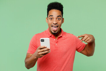 Young smiling happy fun man of African American ethnicity he wears pink t-shirt hold in hand use point finger on mobile cell phone isolated on plain pastel light green background. Lifestyle concept.