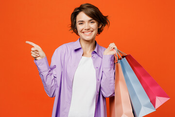 Young happy woman in purple shirt casual clothes hold in hand paper package bags after shopping point aside on area isolated on plain orange color background studio Black Friday sale buy day concept