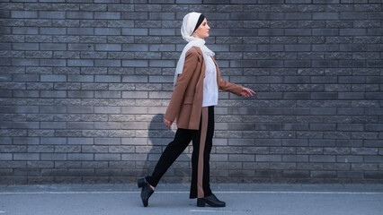 A young woman dressed in a hijab and a business suit walks along a brick wall. 