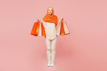 Full body young arabian asian muslim woman wear orange abaya hijab hold shopping package bags lok aside isolated on plain pink background. Black Friday sale buy day, uae middle eastern islam concept.
