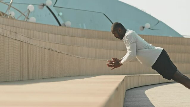 Strong black man in sportswear doing clapping push ups at urban public space outdoors in city while training in morning