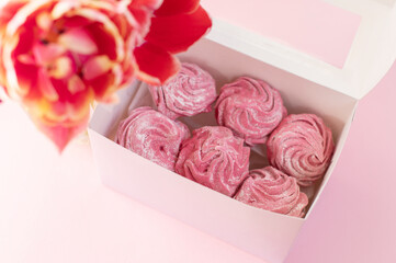 Pink marshmallow lies in a box near a vase with tulips on a pink background