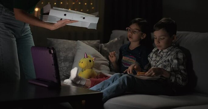 TV night. Kids are using digital tablet for movie night. A woman giving pizza to kids. Children watch TV, eat pizza and relax with online video at night