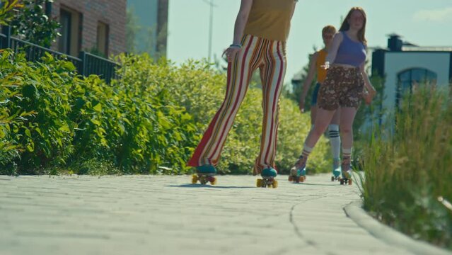 Ground level full shot of three happy gen z girls in bright retro clothes riding on quad skates towards camera outdoor at daytime