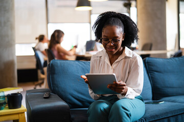 Fototapeta premium Beautiful African businesswoman sitting on sofa in office. Young woman using digital tablet.