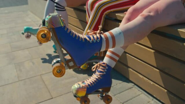 Low angle medium shot of unrecognizable teenage girl sitting on bench wiggling legs in roller skates outside at daytime