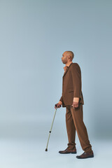 real people, full length view of african american man with myasthenia gravis leaning on walking cane on grey background, dark skinned person in suit, diversity and inclusion