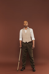 Fototapeta premium real people, bold african american man with myasthenia gravis syndrome standing with walking cane on brown background, standing and looking at camera, diversity and inclusion, physical impairment