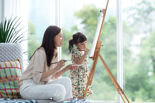 Cute little girl and mom spending good time on weekend coloring on canvas together in living room at home, beautiful green garden in the blurred background, family activity concept