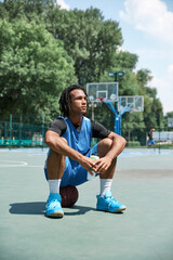 Young man with dreads, athlete in blue uniform sitting on basketball ball, resting after outdoors...