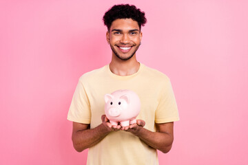 Fototapeta na wymiar Photo of good mood toothy beaming guy with earrings dressed yellow t-shirt hands hold pig money box isolated on pink color background