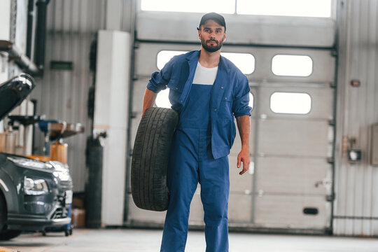 Front View, Standing And Holding Tire. Auto Mechanic Working In Garage. Repair Service