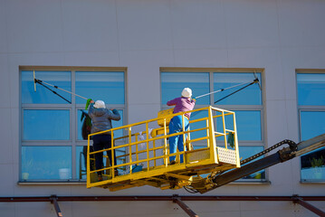 Window cleaner cleaning windows with squeegee, building exterior wet wash. Window cleaning, washing glass windows at height in crane bucket. Workers team washing building on aerial work platform