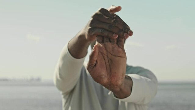 Cropped shot of black male athlete stretching his wrists in front of camera while exercising outdoors on summer day