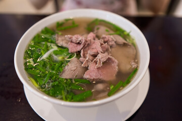 Close up of Vietnamese pho bo spicy broth or soup in white ceramic bowl with rice noodles, beef slices and herbs
