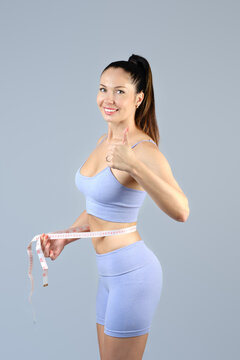 Slimming concept. A slender girl in sports shorts and a tank top shows her thumb, wrapping a measuring tape around her waist. Studio shot on a gray background.