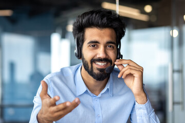 Close-up photo. Portrait of a young Indian man in a headset sitting in front of the camera, smiling, talking, consulting, gesturing with his hands