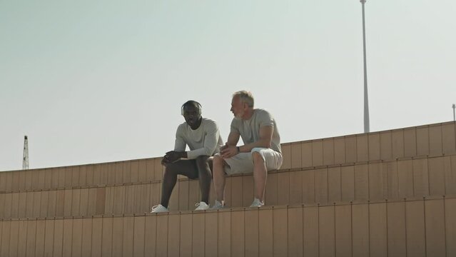 Wide shot of multi-ethnic male friends sitting at urban public space in city, chatting and resting after outdoor workout at daytime