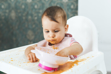 grimy dirty baby 9 months old sits in a highchair and eats complementary foods vegetable puree soup spilled from a plate, the concept of baby food