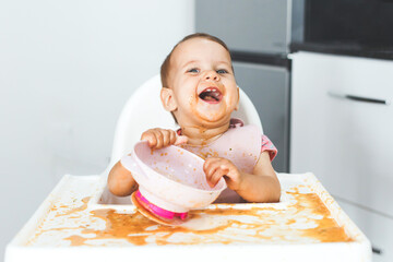 very happy fun cheerful messy baby 9 months old sits in a highchair and eats lure vegetable puree...