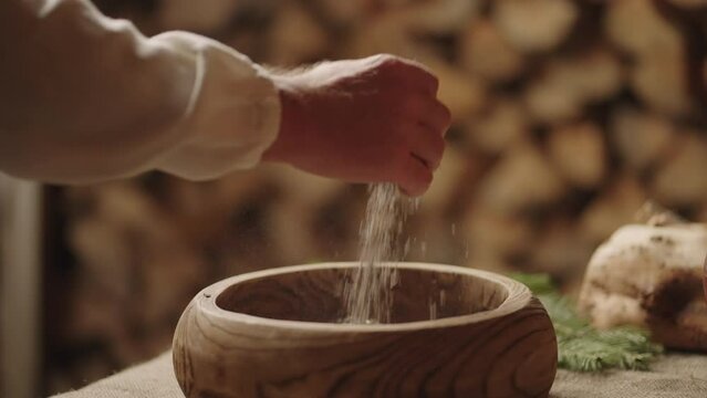 Man Taking Grain From Wooden Bowl On Table In House Of Old Slavic Village, Old Ukrainian Style