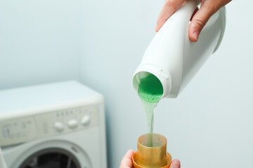 Female hand using or filling detergent in the washing machine. Pour green washing liquid, wash machine blured in background. Female hands pouring liquid laundry detergent into measure cup. - obrazy, fototapety, plakaty