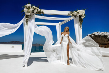 Stylish elegant bride woman in white long fluttering wedding dress is posing in wedding area in Fira in Santorini.