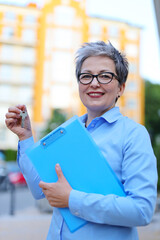 Smiling woman realtor with keys and documents in her hands against the backdrop of a multi-storey building.