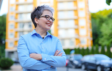 Senior woman realtor with crossed arms against the backdrop of a multi-storey building.