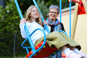 Happy grandmother rolls her granddaughter on a swing in the playground.