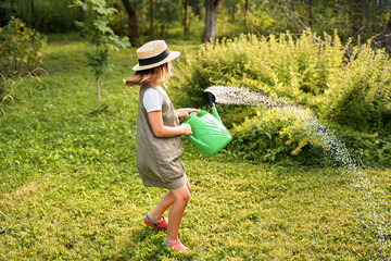 Farmer girl in summer straw hat. Little gardener farming spinning, having fun in garden with big green watering can water fresh grass. Harvest help work. Cultivation healthy organic food, countryside