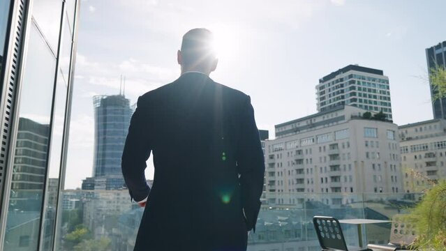 Rear back view following handsome male businessman in suit walks roof top terrace in office building overlooking modern city downtown cityscape with high-rise skyscrapers backlit with bright sunlight