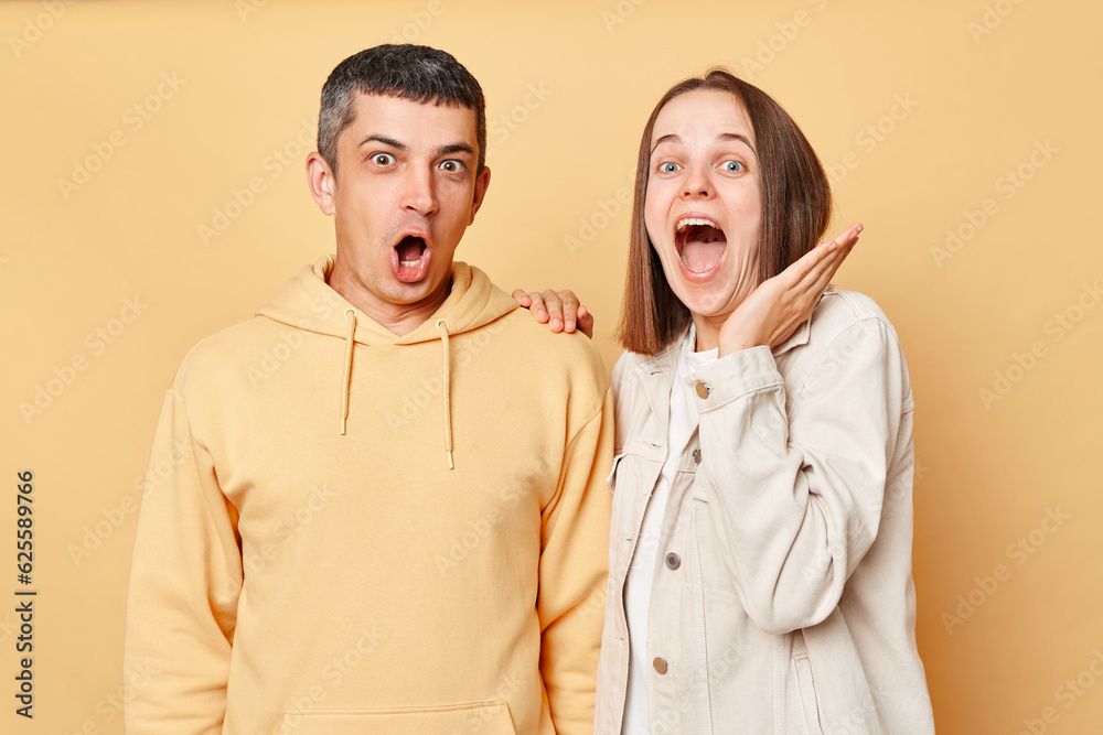 Wall mural Amazed excited woman and man wearing casual style clothing standing isolated over beige background looking at something astonishing with widely opened mouths. - Wall murals