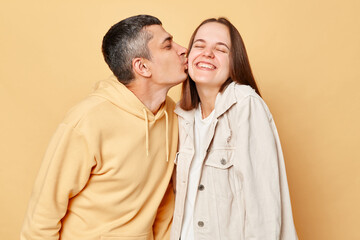 Loving couple woman and man wearing casual style clothing standing isolated over beige background expressing love, husband kissing his wife.