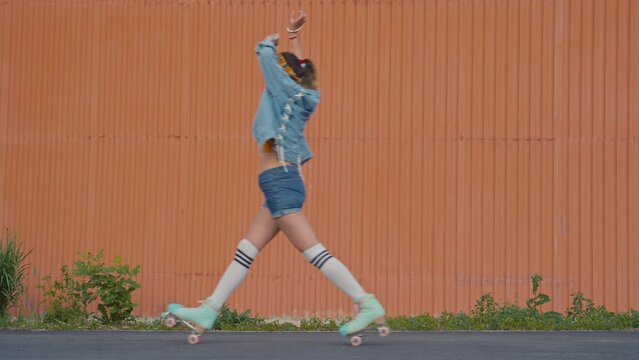 Stable full shot of three bright teenage girls wearing retro clothes and quad skates posing and riding one after another in front of orange wall outside at daytime