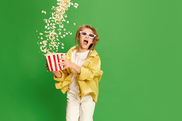 Happy excited little girl in 3d glasses standing with pop corn backet and shouting with delight over green background.