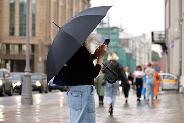 Girl with umbrella and smartphone standing on a street. Rainy weather in summer city