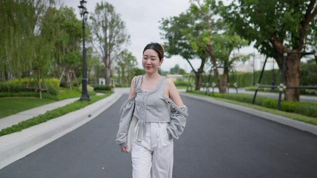 a young Asian person beams with happiness, strolling confidently in the middle of the road, chic grey fashion outfit. The vibrant greenery and urban the joy of stylishly embracing nature's embrace.