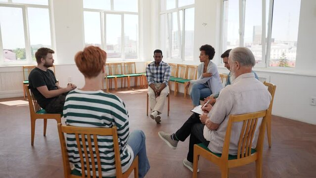 Panning wide shot of African black male drinker talking about alcohol addiction abuse sitting in circle speak to diverse addicted people at group therapy session. Concept of mental health, slowmo.